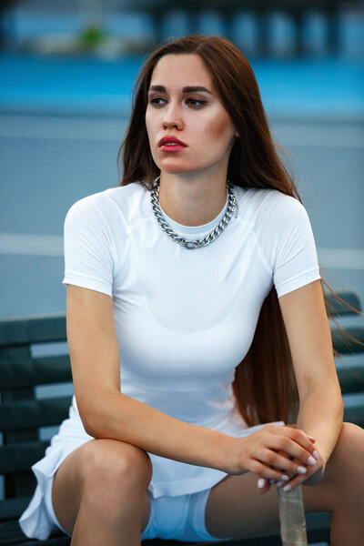 Красивая молодая девушка сидит на скамейке в теннисном корте летом. Портрет модной украинки, позирующей под открытым небом в белой спортивной одежде