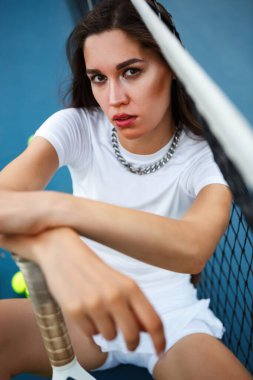 Tenis kortunda poz veren güzel beyaz manken. Elinde raketle oturan şık bir genç kadın.
