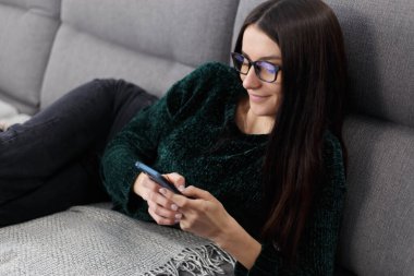 Flört eden genç bir kadın kanepede uzanıyor ve internetten cep telefonuyla mesajlaşıyor. Güzel esmer kadın modern akıllı telefondan mesaj yazıyor.