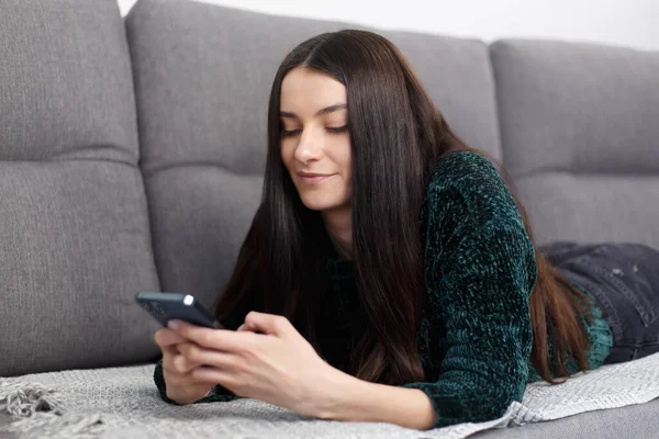 Beautiful brunette woman lying on couch at home and typing a message in smart phone. Cheerful Ukrainian female person in late 20s texting online with a mobile phone