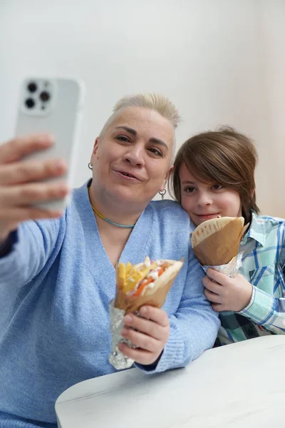 Happy Ukrainian family enjoying lunch meal in a European cafe. Adult white woman taking a selfie together with her son while holding Greek gyros snack in hands