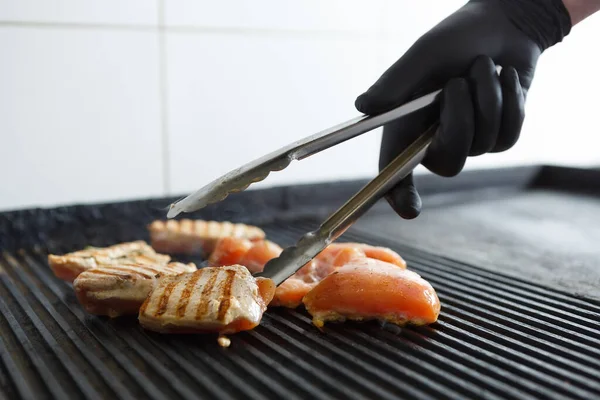 戴着黑色手套的厨师用钳子烤肉 厨师在餐馆厨房里把鸡排放在热锅里烤 — 图库照片