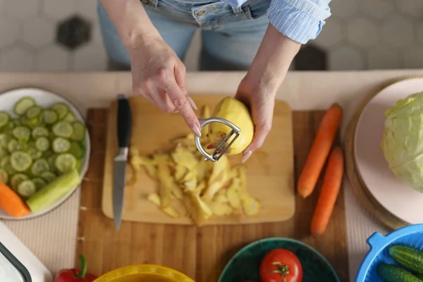 夕食にジャガイモをむいた女性 家で野菜を調理する人 上から撮影 食品用ピーラーツールを使用した女性の手 — ストック写真