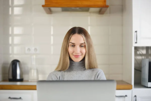 キッチンでコンピュータに取り組んでいる陽気なブロンドの女性 美しい白い女性は自宅でラップトップで遠くの仕事をしています — ストック写真