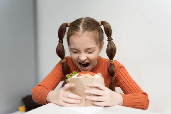 stock image Funny little girl with ponytails biting a sandwich with a grin on her face. Cute hungry kid eating a pita souvlaki snack in a Greek restaurant