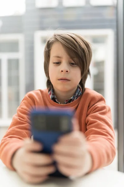 stock image Handsome little boy playing online game on a smart phone. Portrait of pretty white kid sitting in a restaurant and using modern mobile phone