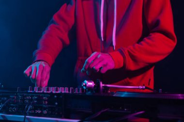 Kulüp DJ 'i, gece kulübündeki ses karıştırıcının sesini ayarlıyor. Kırmızı kapüşonlu profesyonel diskjokeyin partide müzik çalarken yakın plan fotoğrafı.