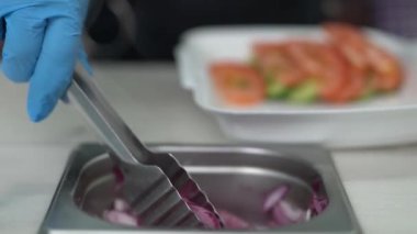 Aşçı dilimlenmiş kırmızı soğanı maşayla birlikte Yunan kalamaki ile birlikte beslenme çantasına koydu. Mutfak işçisi, İspanyol mor soğan halkalarını metal maşayla beslenme çantasına koydu. 
