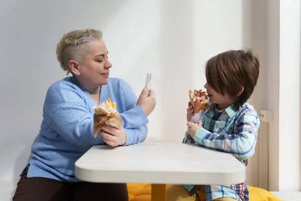 Mother takes a photo of her son eating food in a restaurant. Portrait of short haired woman and 11 year old boy enjoying lunch in a Greek diner