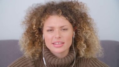 Bir video konuşmasında konuşan ve gülen kulaklıklı genç bir kadının portresi. Kıvırcık saçlı neşeli beyaz kadın kameraya bakıyor ve konuşuyor.