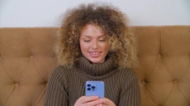 Modern bir akıllı telefondan mesaj yazan kıvırcık saçlı neşeli Ukraynalı kadın. Güzel beyaz kadın yatak odasında oturuyor ve cep telefonuyla mesajlaşıyor.