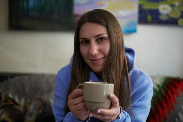 フレンドリーな若いウクライナ人女性がコーヒーを片手にソファに座っています カフェで熱い飲み物を飲むかなり白いブルネットの女性の肖像画 — ストック写真
