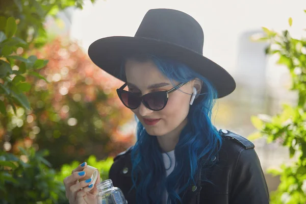 一个漂亮多样的人的画像 他的头发染成蓝色 喝着玻璃瓶里的清水 迷人的白色女性 戴着帽子和太阳镜 用无线耳机听音乐 — 图库照片