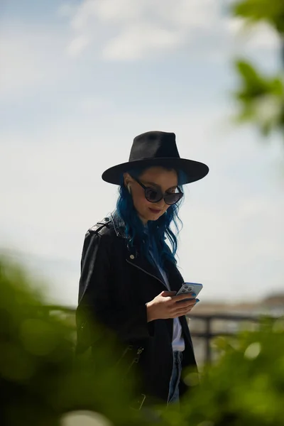 时尚多样的女性与染成蓝色的头发使用智能手机户外 在城市屋顶花园用手机描绘一个戴着嬉皮士帽和皮夹克的时髦年轻女子 — 图库照片