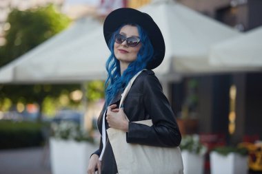 Boyalı mavi saçlı, güzel bir model. Şehir sokaklarında poz veriyor. Omuz çantasına çanta takıyor. Hippi şapkalı ve güneş gözlüklü çekici beyaz kadın dışarıda yürüyor.