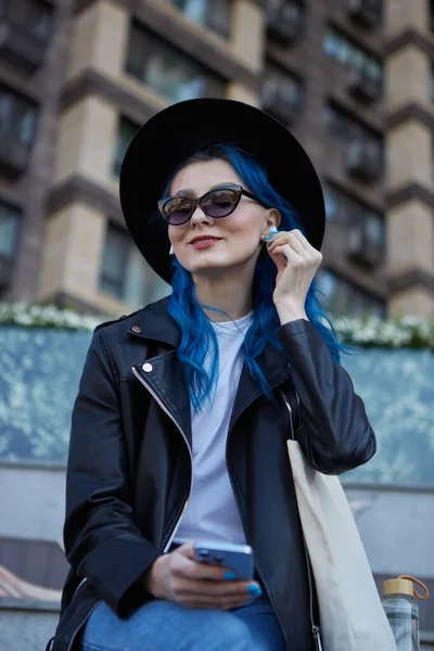 拥有一头染发的快乐而多姿多彩的女人喜欢音乐流线型的无线耳机和智能手机应用 市中心戴着嬉皮士帽和太阳镜的漂亮女性画像 — 图库照片