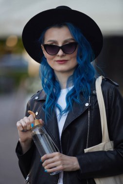 Yeniden kullanılabilir cam bir su şişesini elinde tutan ve son moda bir çanta taşıyan şık bir kadının portresi. Şehirde yürüyen parlak mavi saçlı güzel bir genç kadın.