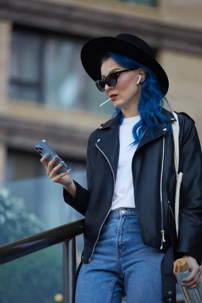 漂亮多样的女性在户外使用智能手机 用无线耳机听音乐 吃棒棒糖 一个染成蓝色头发的时尚白人妇女的画像 — 图库照片