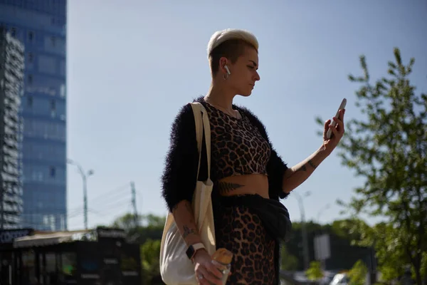 スタイリッシュな多様な女性が肩にヒョウ柄の服やトートバッグを着ている 無線ヘッドフォンで音楽を聴き 携帯電話を屋外で使用する短い髪の女性の肖像画 — ストック写真