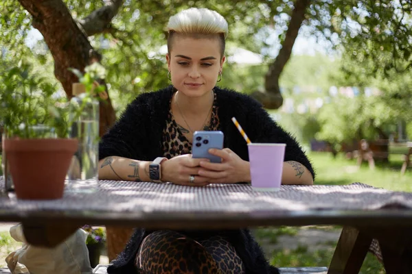 美丽多样的女人 短发坐在一个舒适的绿色花园的木制桌子后面 一个漂亮的白人女性在户外用智能手机输入信息的肖像 — 图库照片