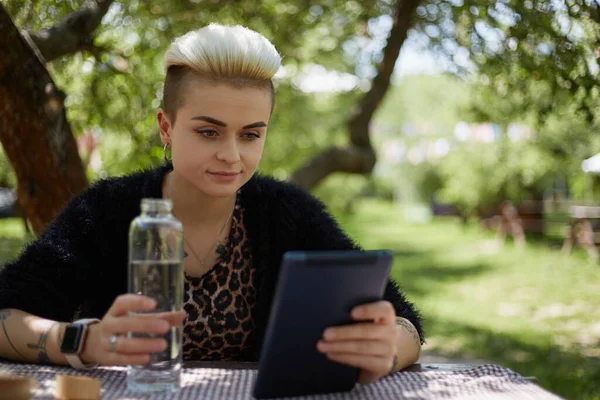美丽的年轻女子从玻璃瓶中喝水 并在一个绿色的公园里用平板电脑阅读电子书 时尚的汤姆男孩的画像 用淡水给自己浇水的女孩 — 图库照片