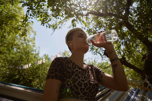 多様な女性がガラス瓶の屋外から水を飲む ハンモックに座っている間 日当たりの良い公園で新鮮な水を飲むスタイリッシュな短い髪の女性の肖像画 サステナビリティと多様性の概念 — ストック写真