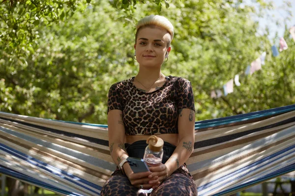 手に水のガラスボトルを持つ公園でハンモックに座って短い髪を持つフレンドリーな若い入れ墨の女性 緑の庭にポーズをとる陽気で多様な女性 — ストック写真