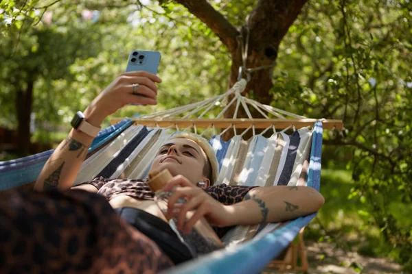 年轻而多样化的女人 短发躺在公园的吊床上 在智能手机上浏览手机应用程序 夏天的周末 文雅的纹身女人们在一个绿色的花园里悠闲自在地玩耍 — 图库照片