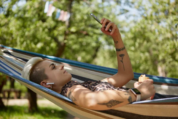 年轻多样的女性躺在一个绿色花园的吊床上 用无线耳机听音乐 漂亮的纹身妇女 留着短发 手握智能手机 在吊床上悠闲自在 — 图库照片