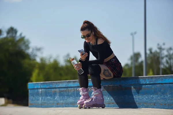 年轻的女滑板手坐在滑板上 手里拿着玻璃瓶水和智能手机 滑雪场里穿着现代紫色内联滑板的轮滑手 — 图库照片