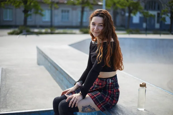 可爱的红头发滑板手坐在一个城市的滑板公园里 手里拿着玻璃瓶水 年轻漂亮的成年女子准备在室外混凝土公园滑冰 — 图库照片