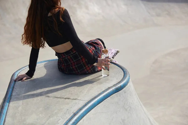 滑板女孩坐在一个滑板公园的混凝土池子上 手里拿着玻璃瓶水 穿着内联滑冰鞋的难以辨认的女性骑完车后会在一个滑冰场里发冷 — 图库照片
