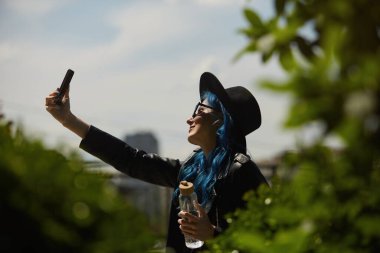 Mavi saçlı çeşitli kadınlar çatı bahçesinde cam bir su şişesiyle selfie çekiyorlar. Siyah deri ceketli ve hippi şapkalı şık genç bir kadın açık havada selfie çekiyor. 