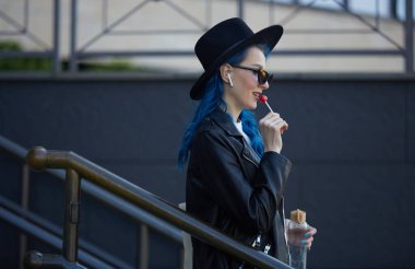 Mavi saçlı, şık bir kadın caddede yürüyor ve lolipop şekeri yalıyor. Hippi şapkalı neşeli bir kadın yürüyüş sırasında müzik dinliyor.