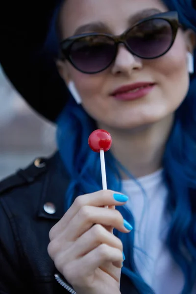 时尚的年轻女性 染成蓝色的头发 在户外吃糖果 戴着太阳镜 听音乐 舔棒棒糖的漂亮女人 — 图库照片