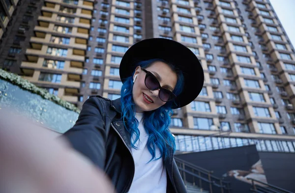 現代の街の中心部にある高層ビルでは ハイスタイリッシュで多様な女性の帽子とサングラスがセルフィーを取っています 美しいです若いです女性で染めた青い髪のポーズのために自画撮り写真付き笑顔 — ストック写真