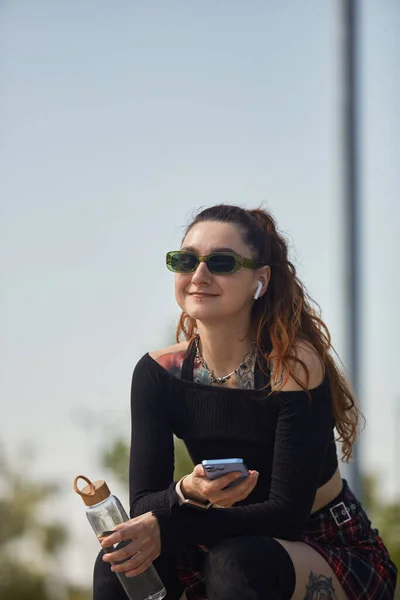 戴着太阳镜的年轻漂亮的女人坐在一个滑板公园里 听着无线耳机里的音乐 喝着可重复使用的玻璃瓶里的淡水 — 图库照片
