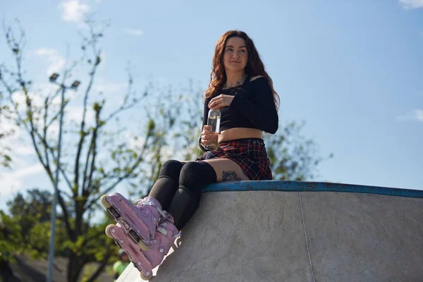 ローラーブレーダーの女性は コンクリートスケートプールのランプの上に座って ガラスボトルから水を飲んでいます クールな若いですスケートパークで現代的な積極的なインラインスケートを身に着けている — ストック写真