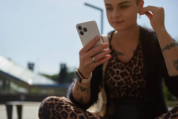 穿着豹衣的年轻女性 一边用耳机听音乐 一边在现代智能手机上浏览应用程序 坐在长椅上使用手机的纹身女性 — 图库照片