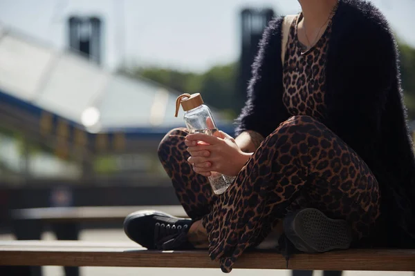 ヒョウ柄の服の若い女性が手に新鮮な水のガラスボトルとベンチに座って印刷します 多様性と持続可能性の概念 — ストック写真