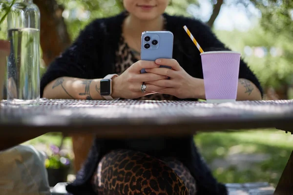 快乐的年轻女性坐在一棵树下的野餐桌上 使用智能手机 喝着可回收纸杯中的鸡尾酒 美丽多样的女人在一个绿色的花园放松周末 — 图库照片