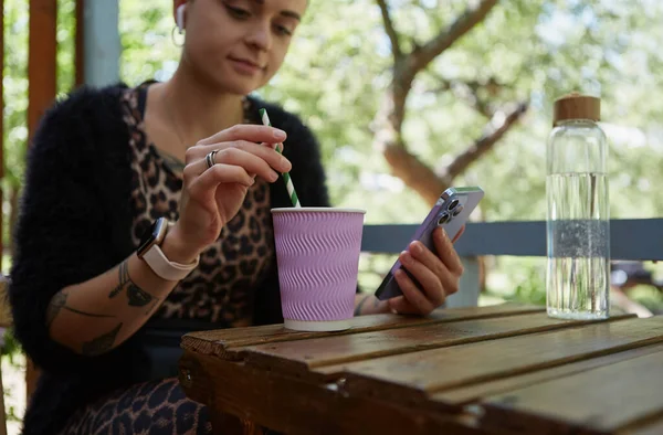 年轻女子坐在室外咖啡馆里 一边浏览智能手机 一边喝着紫色纸杯里的鸡尾酒 — 图库照片