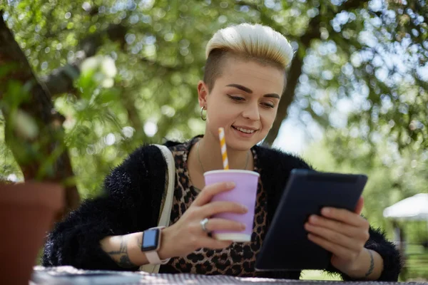 一个快乐而多样化的女人坐在一个绿色的公园里一张花哨的桌子后面 浏览着平板电脑 喝着纸杯里的鸡尾酒 一个有着短发的漂亮的白色女性的画像 — 图库照片