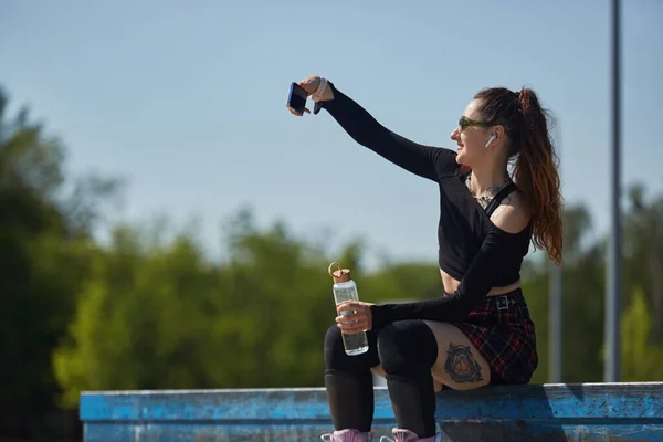 快乐的滚筒刮胡刀女拿着一个生态玻璃瓶自拍 一个滑板手坐在滑板上 微笑着摆出一张自拍照片的肖像 — 图库照片