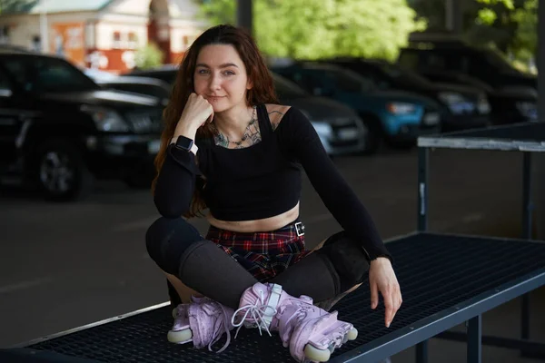 在线滑板女选手坐在一个滑板公园里 脚上有现代的攻击性轮滑刀片 一个有纹身胸部 头戴溜冰鞋和护膝垫的年轻漂亮女子的画像 — 图库照片