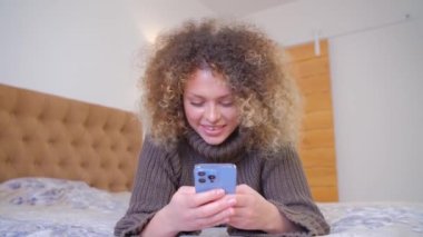 Yatak odasındaki yatakta kıvırcık saçlı ve akıllı bir telefonla internetten iletişim kuran mutlu genç bir kadın. Neşeli beyaz kadın iletişim için cep telefonu kullanıyor.