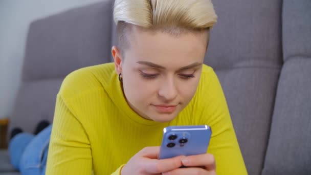 美丽多样的女人 短短的漂白头发躺在沙发上 用智能手机打字 用现代蓝色手机刻画漂亮的汤姆男孩的女性形象 — 图库视频影像