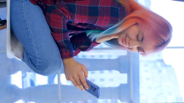 坐在窗边的椅子上 在智能手机上浏览手机应用程序 有着彩虹色头发的快乐而多样化的女人 — 图库视频影像