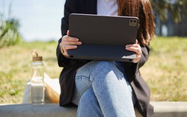 Güneşli bir parkta elinde tablet bilgisayar ve cam bir şişe suyla oturan genç bir kadın. Serbest çalışan kişi modern taşınabilir bir cihaz üzerinde çalışıyor.
