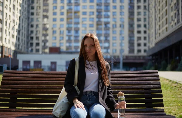 高層ビルの近くの街の通りにベンチに座って長い茶色の髪を持つ美しいウクライナの女性 20代のかなり若い女性の肖像画手にガラス水ボトルでポーズ — ストック写真